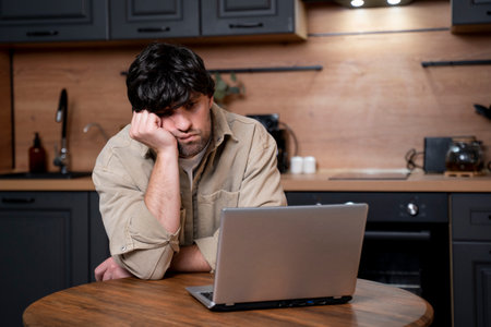 Stressed man working on laptop in the kitchen at home. Distance work concept. A tired brunette man is working on a laptop in the kitchen.の写真素材