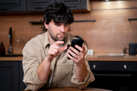 A young brunette man dressed in a beige shirt uses a smartphone sitting at a table in the kitchen.の写真素材