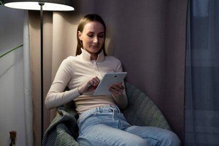 A happy young woman is resting at home in a comfortable chair using a modern tablet gadget. A smiling woman enjoys a cozy home weekend in the living room while browsing a tabletの写真素材