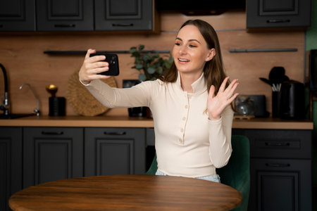 A young brunette woman makes a video call from her smartphone while sitting in the kitchen at home.の写真素材