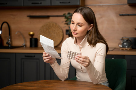 Young brunette woman checks the prescription and dosage of the medicine. A woman reads medical instructions before taking medication while sitting at a table in the kitchen.の写真素材