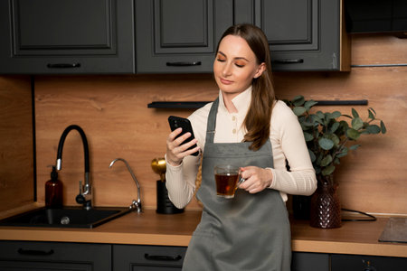 Happy beautiful woman in an apron using a mobile phone while drinking coffee in a modern kitchen.の写真素材