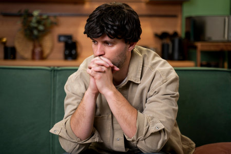 I feel stressed and upset. A disappointed handsome young man touches his head, worried about breaking up or losing his job, sitting at home on a green sofa.の写真素材