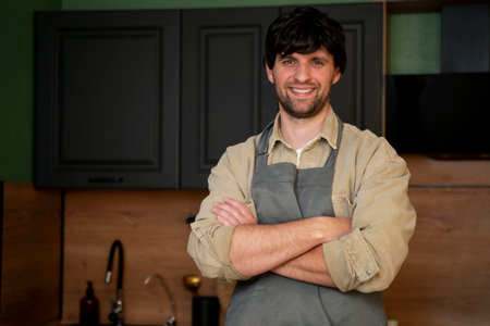 Handsome smiling young man in an apron looking at the camera. Portrait of a happy laid-back guy in an apron standing in the kitchen.の写真素材