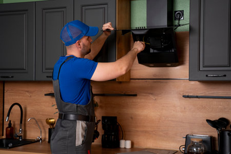 A handyman in uniform, installs or repairs a kitchen hood, replaces the filter in the hood. Construction, maintenance and repair concept.の写真素材