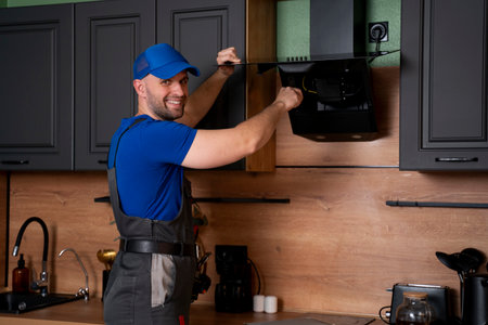 A handyman in uniform, installs or repairs a kitchen hood, replaces the filter in the hood. Construction, maintenance and repair concept.の写真素材