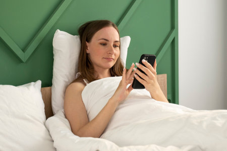Portrait of a beautiful young brunette woman using a smart mobile phone on a bed with a white pillow-blanket in the bedroom interior.の写真素材