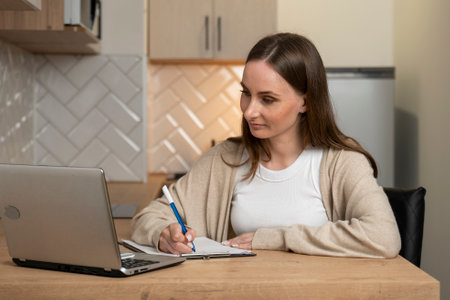 Pretty young girl sitting at a desktop computer, holding an open notebook, writing in a home office in the kitchen.の写真素材