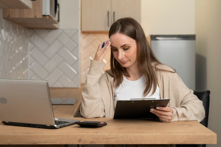 Serious young woman calculating finances, household expenses, working with project statistics using a laptop and a calculator, sitting at home in the kitchen.の写真素材