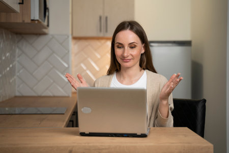 A positive student, smiling, sitting at a table with a laptop in the kitchen, studying at home and waving at the screen, watch an online webinar.の写真素材