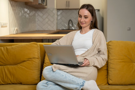 Young woman in casual clothes is sitting on a yellow sofa using a laptop, having fun on social networks, relaxing, smiling and laughing.A female freelancer engaged in online sales marketingの写真素材