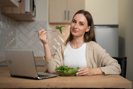 A woman is sitting at a table in the kitchen and enjoying a nutritious salad for lunch and using a laptop.の写真素材