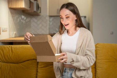Fast delivery. A happy woman has received a parcel, unpacks a cardboard box, sits at home on a yellow sofa. A customer excited about her purchase in an online store.の写真素材