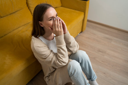 Depressed woman is sitting on the floor in the living room near a yellow sofa.の写真素材