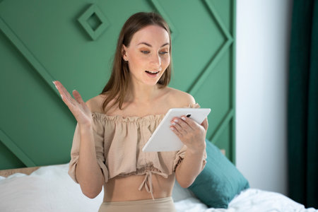 Beautiful young brunette woman smiles and uses her tablet on the bed.の写真素材
