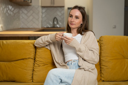 Pretty dreamy young woman is sitting on a yellow sofa, and drinking coffee in the living room. Cozy scene, the concept of a cozy home.の写真素材