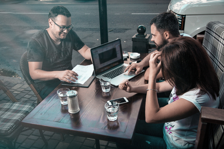 Side view and top. The company of three businessmen are discussing at the table in coffee, writing in a notebook, looking at a laptop, satkan with water, car keys, magazines, a smartphone, online transactions.の写真素材