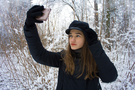 Fashion pretty beautiful attractive young teen girl with long blonde hair with gray green eyes makes selfie self portrait on smartphone on natural gentle blurred blue and purple romantic background in frosty snowy winter forest.の写真素材
