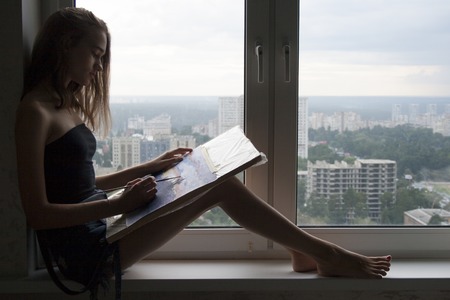 Silhouette of young woman is drawing a watercolor painting. The female artist paints watercolour landscape. Teen girl with blond hair is sitting on the window sill with brush in hand. Creative conceptの写真素材