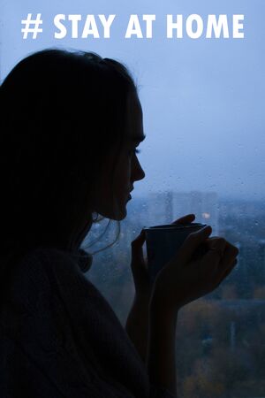 Stay at home virus quarantine background. Coronavirus backdrop with lettering, silhouette of young beautiful woman with cup of tea near window. Pandemic protection concept.の写真素材