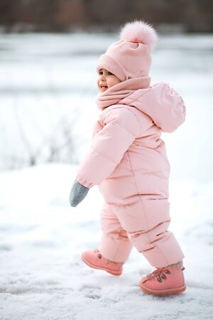 A beautiful young girl wearing a pink jumpsuit running in a snowy winter park. Kid plays and jumps in a snowy forestの写真素材