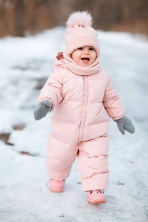 A beautiful young girl wearing a pink jumpsuit running in a snowy winter park. Kid plays and jumps in a snowy forestの写真素材