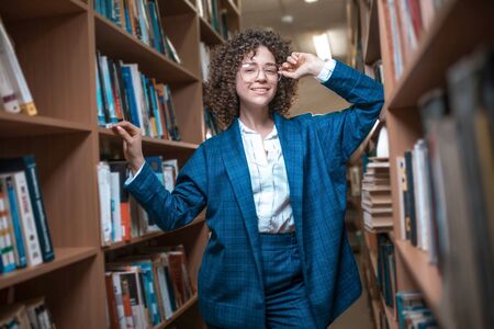 Young beautiful curly girl in glasses and a blue suit is standing in the library. Student Studyの写真素材