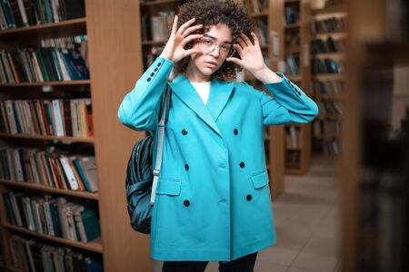Young beautiful curly girl in glasses and a blue suit is standing in the library. Student Study
