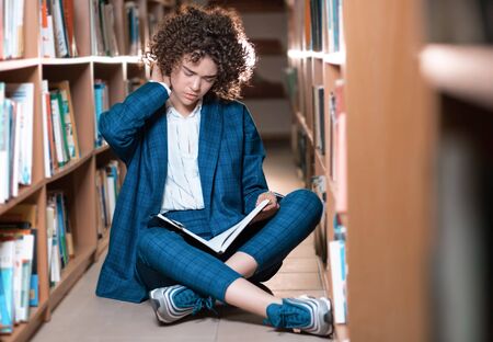 Young beautiful curly girl in glasses and blue suit sitting with books in the library. Student Study