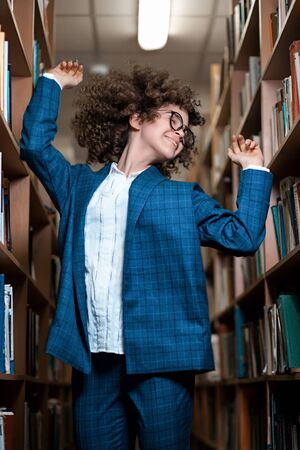 Young beautiful curly girl in glasses and a blue suit is standing in the library. Student Study