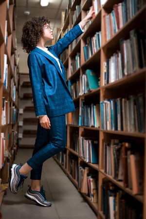 Young beautiful curly girl in glasses and a blue suit is standing in the library. Student Study