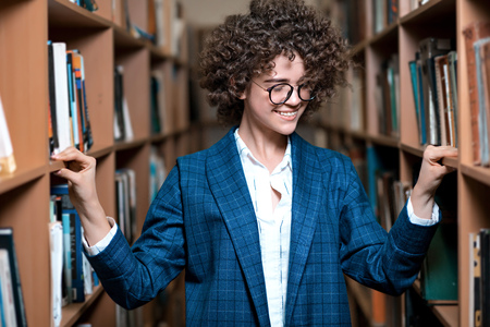 Young beautiful curly girl in glasses and a blue suit is standing in the library. Student Studyの写真素材