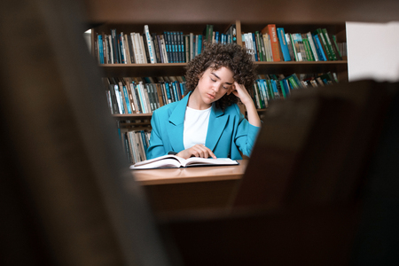 Young beautiful curly girl in glasses and blue suit sitting with books in the library. Student Study
