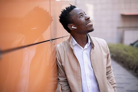 handsome black young man in a jacket and white shirt is listening to music with headphones on an orange backgroundの写真素材