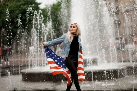 beautiful young woman blonde on a background of the city stands with the American flagの写真素材