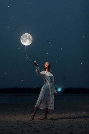 Young attractive girl in a long white dress on the beach holds the moon on a rope. Art photographyの写真素材