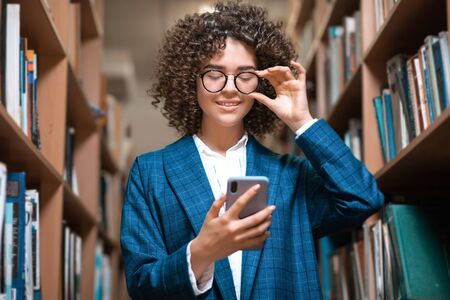 Young beautiful curly girl in glasses and a blue suit is standing in the library. Student Studyの写真素材