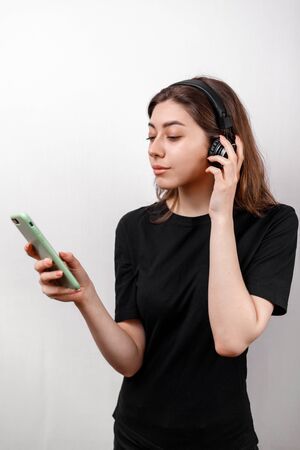 young brunette woman in a black t-shirt on a white background listens to music with a phone. isolatedの写真素材