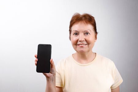 Senior woman shows a phone on a white background in a light T-shirt. place for text. isolatedの写真素材