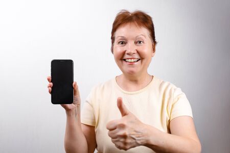 Senior woman shows a phone on a white background in a light T-shirt. place for text. isolatedの写真素材