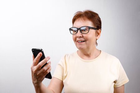 Senior woman in glasses looks at the phone on a white background in a light T-shirt. place for text....
