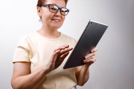 Senior woman in glasses uses a tablet on a white background in a light T-shirt. place for text. isolatedの写真素材