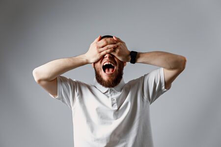 Portrait of a happy young man in a white T-shirt laughing and closing his eyes with his hands on a...