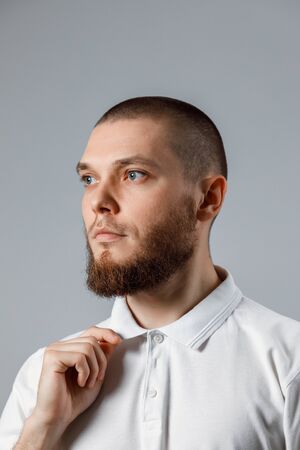 Portrait of a handsome young man in a white T-shirt straightens his collar looks away on a gray...