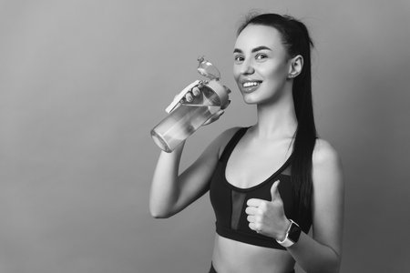 beautiful young brunette woman in sportswear stands with a bottle of water showing thumbs up on a...