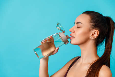 close-up portrait of a beautiful young brunette woman in a sportswear drinks water from a bottle on a blue background. space for textの写真素材