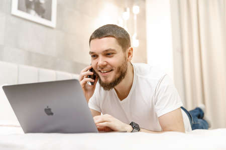 young man at home on the couch talking on the phone and looking at the laptop.