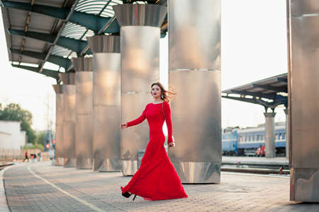 beautiful young woman in a long red dress stands on the platform of the station near the railway.の写真素材
