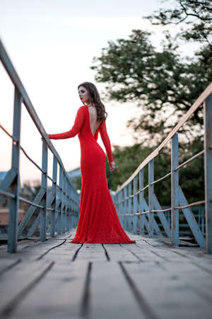 portrait of a beautiful young woman in a long red dress stands on the bridge.の写真素材