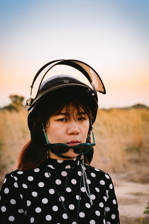 The woman wear polka dot with motorcycle helmet and sunglasses posting near meadow with twilight time.の写真素材
